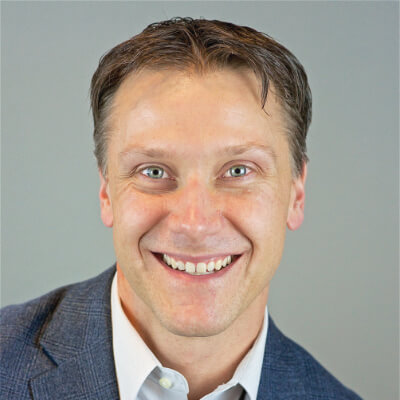 Ryan Brager, Eaton. A smiling man with light brown hair, wearing a blue blazer and white collared shirt, poses in front of a plain, light gray background.