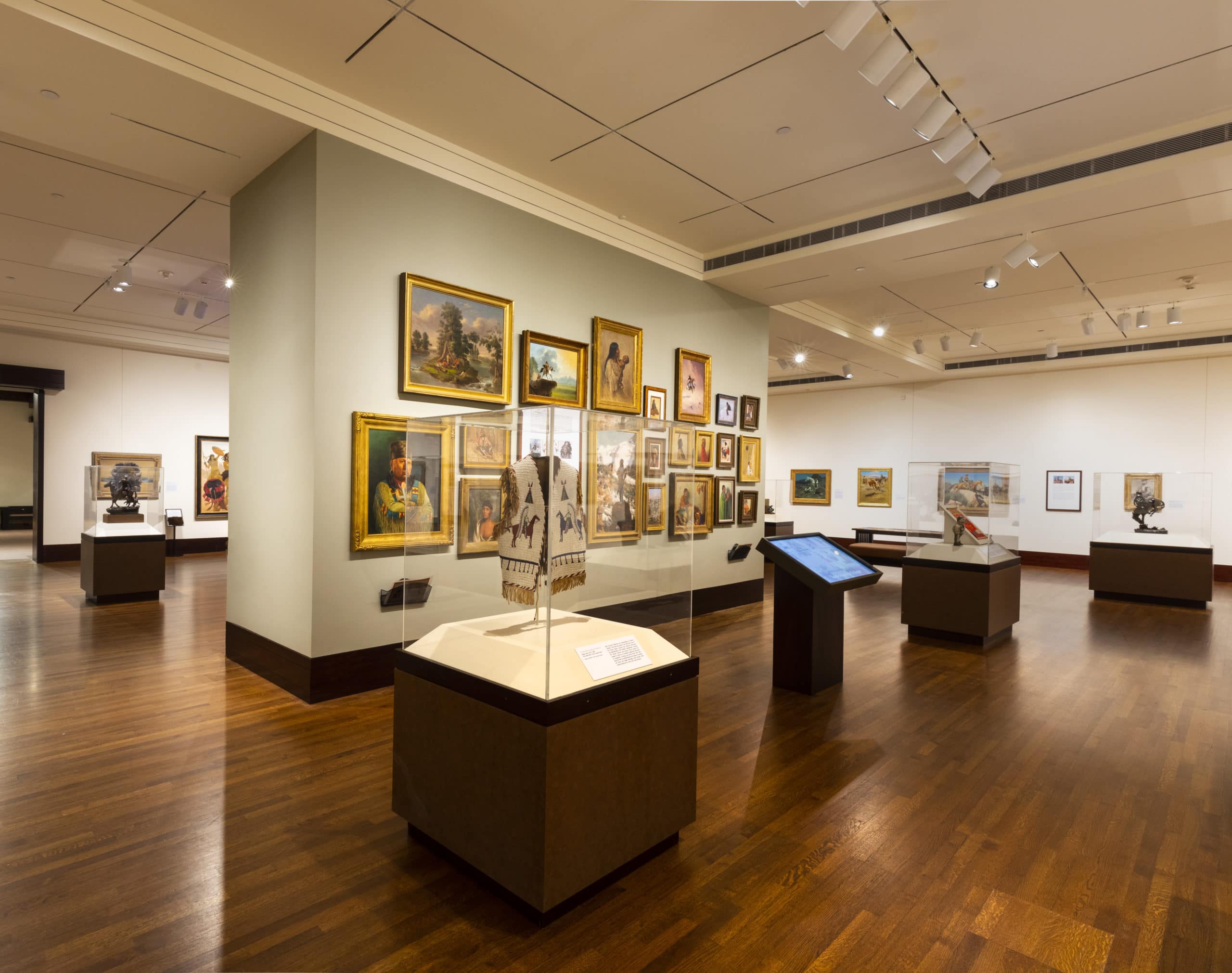 A spacious art gallery with wooden floors, featuring framed paintings on pale walls and display cases with sculptures and artifacts. Track lighting illuminates the artworks, and digital information kiosks are present throughout the room.