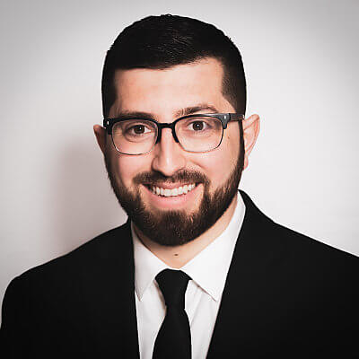 A man with short dark hair, a beard, and glasses is smiling. He is wearing a black suit, white shirt, and black tie, and is posed against a plain, light-colored background.
