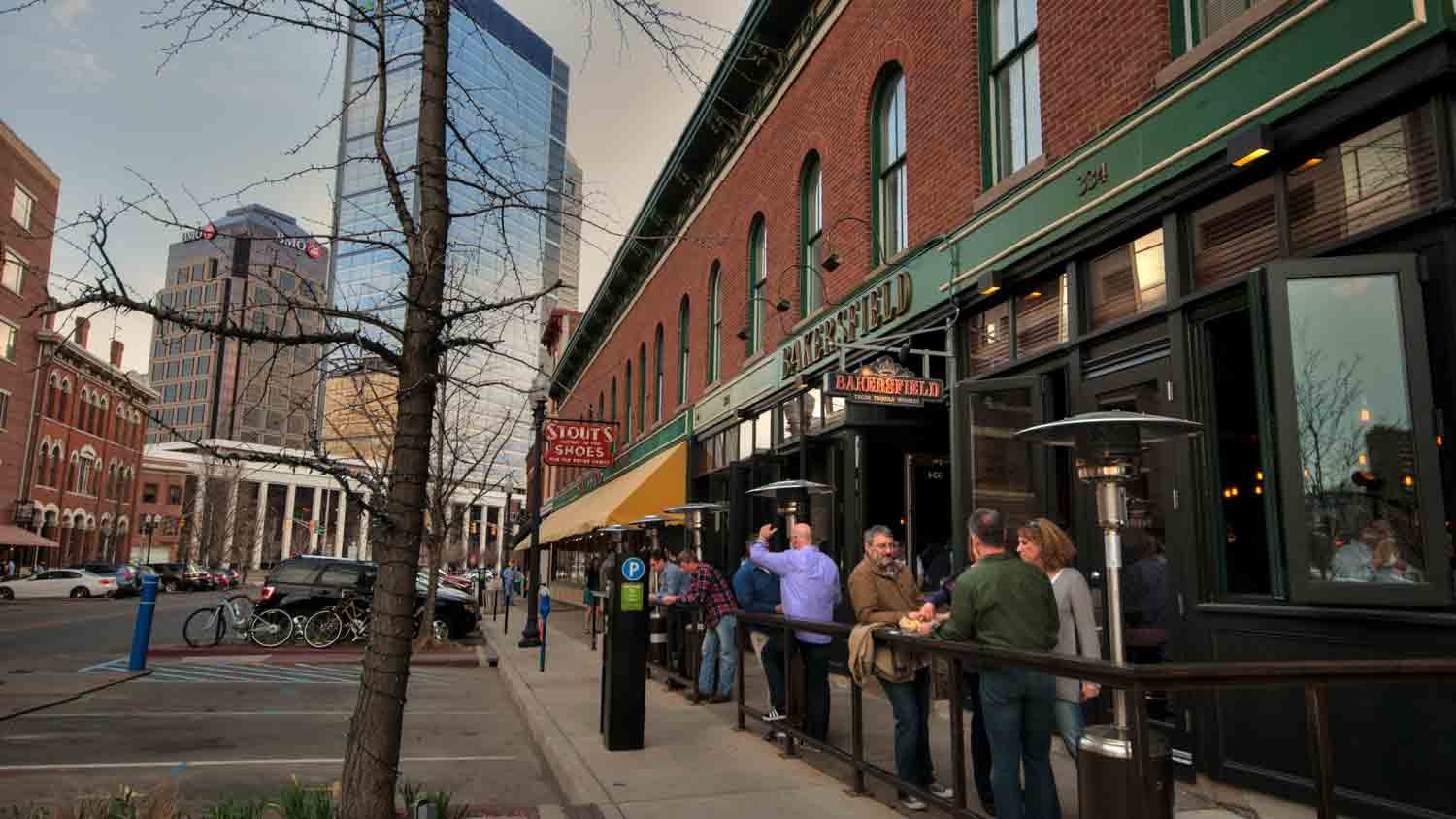 People wait in line outside a brick restaurant with green awnings on a city street. Tall modern buildings and parked bikes are visible in the background on an overcast day.