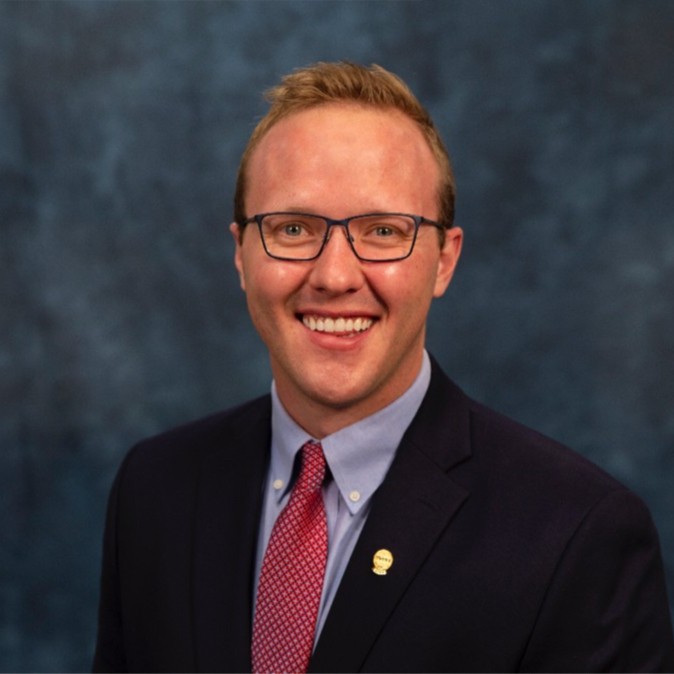 A smiling man with short blond hair and glasses, wearing a dark suit, light blue shirt, and red patterned tie, poses in front of a mottled blue background.