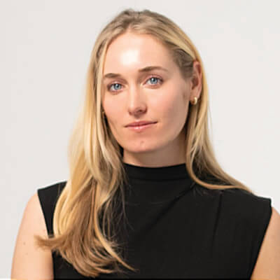 A young woman with long blonde hair, wearing a navy blue sweater over a white collared shirt, smiles at the camera with her arms crossed in front of her. The background is softly blurred.