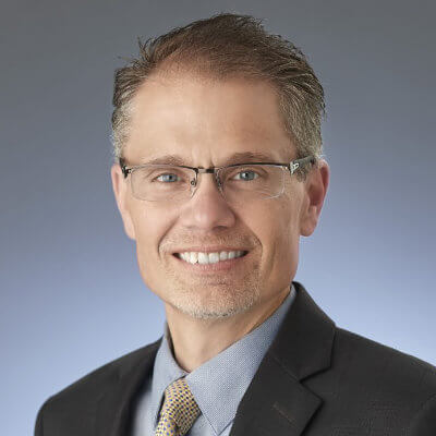 A smiling man wearing glasses, a dark suit, a blue shirt, and a patterned tie, posing in front of a plain, gradient gray background.