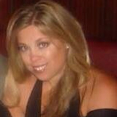 A woman with long blonde hair and light skin smiles at the camera. She is wearing a black top and is seated indoors with a dark background.