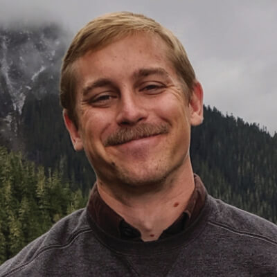 A man with light brown hair and a mustache smiles outdoors, standing in front of a backdrop of evergreen trees and misty mountains under a cloudy sky.