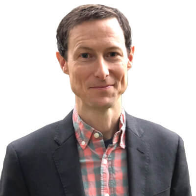 A man with short brown hair wearing a dark blazer over a pink and blue plaid shirt, smiling slightly, standing against a plain white background.