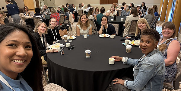 A group of women from Women in Load Flexibility sit around a round table, smiling at the camera with coffee cups and plates of food, in a large, bright conference room filled with other people and tables.