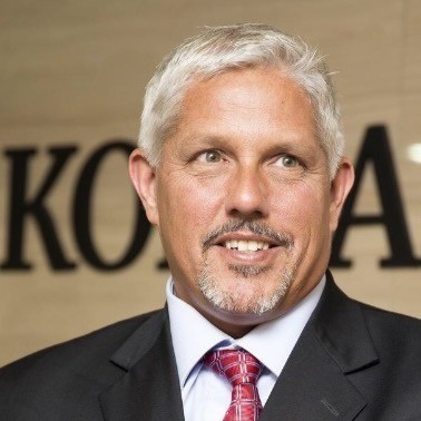 Aaron Breidenbaugh, Cpower. A middle-aged man with short gray hair and a goatee, wearing a dark suit, white shirt, and red patterned tie, stands in front of a blurred sign with black letters. He is smiling and looking slightly to the side.