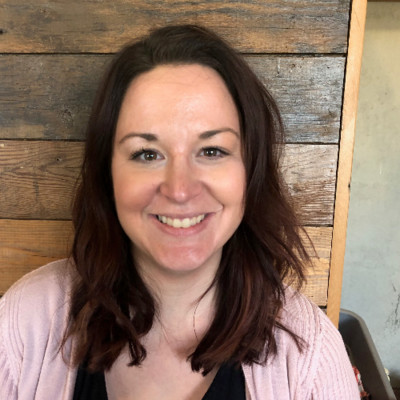 Beth Glynn, Cascade Energy. A woman with shoulder-length brown hair and a pink cardigan smiles at the camera while sitting in front of a wooden wall.