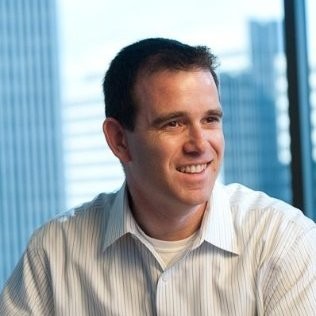 A man with short dark hair, wearing a light striped button-up shirt, smiles and looks to the side in an office with large windows and tall buildings in the background.