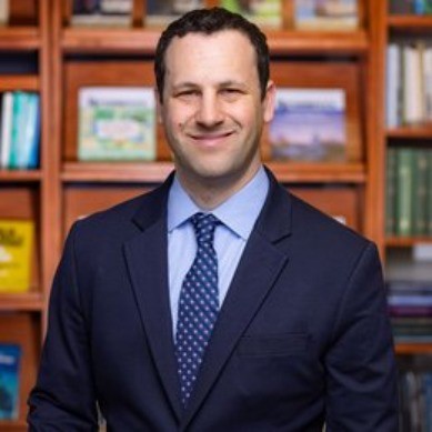 Brad Harris, WeaveGrid. A man in a suit and tie is smiling while standing in front of a bookshelf filled with books.