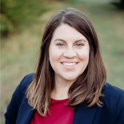 A woman with long brown hair, wearing a navy blazer over a red top, smiles outdoors with greenery and a blurred background behind her.