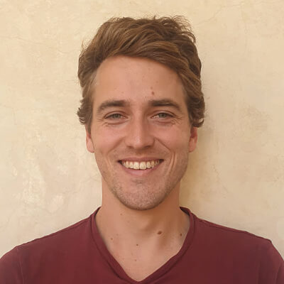 A young man with light brown hair and fair skin smiles at the camera. He is wearing a maroon shirt and stands in front of a beige textured wall.