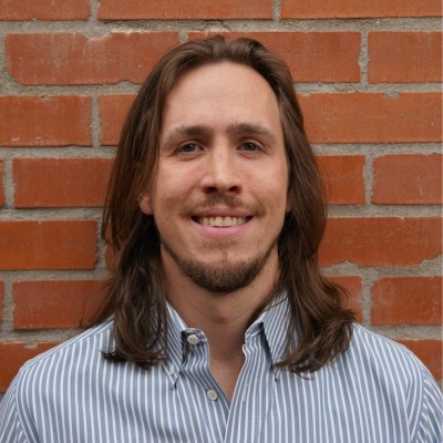 A man with long brown hair and a beard, wearing a blue and white striped button-up shirt, smiles while standing in front of a brick wall.