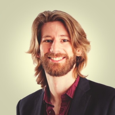 David Pudleiner, ICF. A man with long, light brown hair and a beard is smiling. He is wearing a dark blazer over a burgundy shirt. The background is a plain, light neutral color.