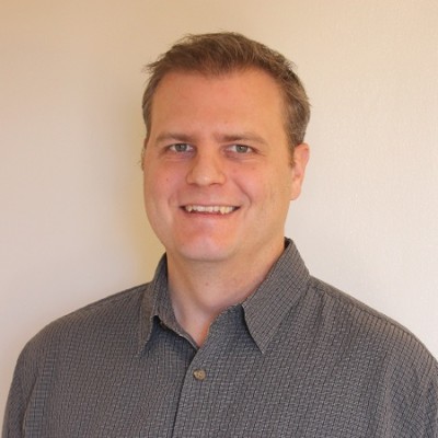 A man with short light brown hair wearing a gray checkered button-up shirt smiles at the camera in front of a plain light-colored background.