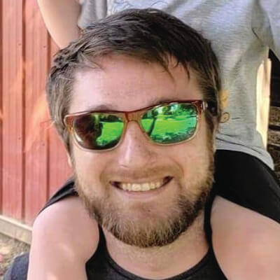 Eric Ricucci, Baltimore Gas and Electric. A smiling man with a beard and sunglasses carries a child on his shoulders. The child’s legs and part of their arms are visible, but their face is out of frame. There is a red wooden wall in the background.