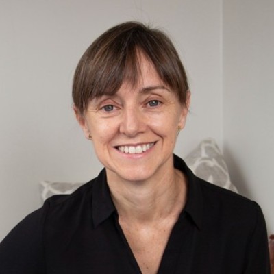 A woman with short brown hair and bangs, wearing a black collared shirt, smiles at the camera while seated indoors against a light-colored background.