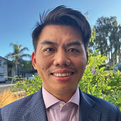 John Hernandez, Pacific Gas and Electric Company. A man in a checked suit jacket and light pink shirt smiles outdoors, with greenery and trees in the background on a sunny day.