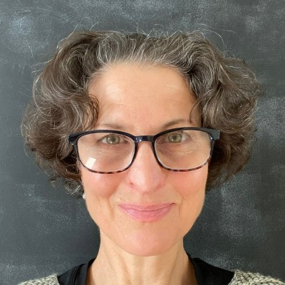 Julie Michals, NASEO. A woman with short, curly gray hair and glasses smiles warmly at the camera. She is standing in front of a dark chalkboard background and wearing a light-colored sweater.