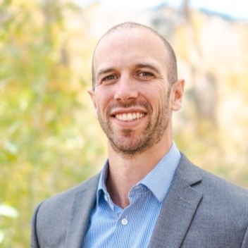 Kevin Brehm, RMI. A man with a shaved head and trimmed beard, wearing a gray blazer and blue shirt, smiles outdoors in front of a blurred background with green and yellow foliage.