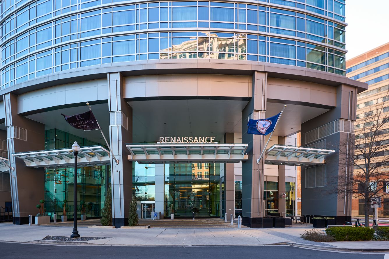 The main entrance of a modern glass building with Renaissance signage above the doorway, two flags, and reflections of surrounding buildings in the windows.