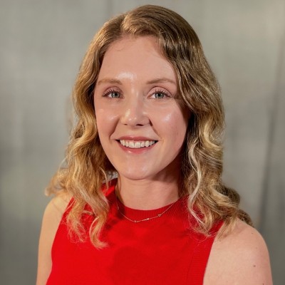 Sarah McCarville, MidAmerican Energy Company. A woman with wavy blonde hair is smiling at the camera. She is wearing a sleeveless red top and a thin necklace, standing in front of a neutral, softly blurred background.