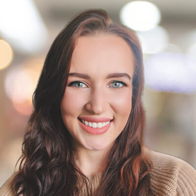 Victoria Cooper, AES Indiana. A young woman with long brown hair and blue eyes smiles at the camera, wearing a beige sweater. The background is softly blurred with warm, circular lights.