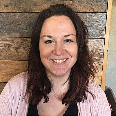 Beth Glynn, Cascade Energy. A woman with shoulder-length brown hair is smiling at the camera. She is wearing a light pink cardigan and a black top, sitting in front of a wooden wall background.