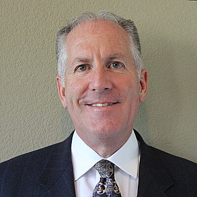 Christian Lenci, Linde. A middle-aged man with short gray hair is wearing a dark suit, white dress shirt, and a patterned tie, standing in front of a plain light-colored wall, smiling at the camera.
