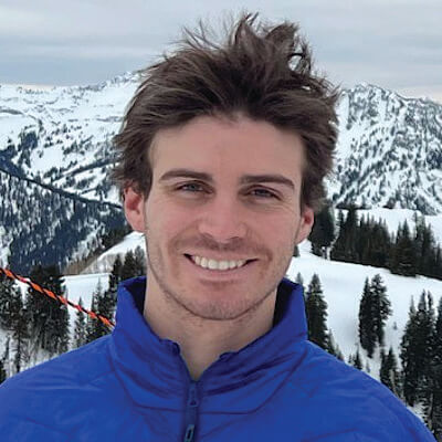 A young man with tousled brown hair and a blue jacket smiles outdoors, with snow-covered mountains and evergreen trees in the background.