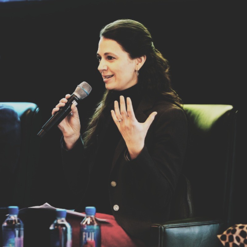 Cristina Botero, ComEd. A woman with long dark hair sits in a chair, speaking into a microphone and gesturing with her hand. She is wearing a dark blazer and appears to be participating in a panel discussion. Bottled water is on the table in front of her.