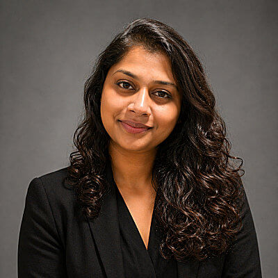 Devi Mohan, Con Edison. A woman with long, wavy dark hair wearing a black blazer poses in front of a plain gray background, smiling softly at the camera.