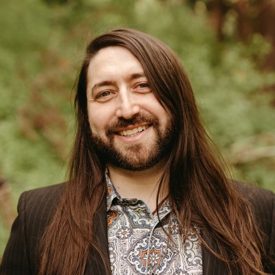 Jason Trager, Plentiful.ai. A person with long brown hair and a beard smiles outdoors. They are wearing a patterned shirt and a dark pinstripe blazer, with a blurred green natural background.
