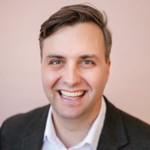Jeremy Hutchinson, Xcel Energy. A man with short brown hair, wearing a dark blazer and white shirt, is smiling broadly at the camera against a plain light pink background.
