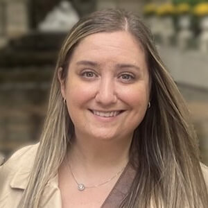Katie Davis, ComEd. A woman with long, straight, light brown hair smiles at the camera outdoors. She is wearing a beige jacket and a necklace. The background is blurred, showing greenery and a stone structure.