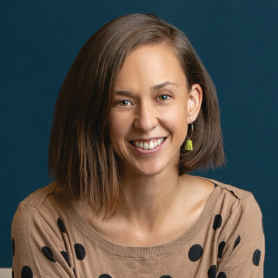 Laura Tajima, Renew Home. A woman with straight brown hair and an asymmetrical bob smiles at the camera. She is wearing a tan sweater with black polka dots and a single yellow earring, against a dark blue background.
