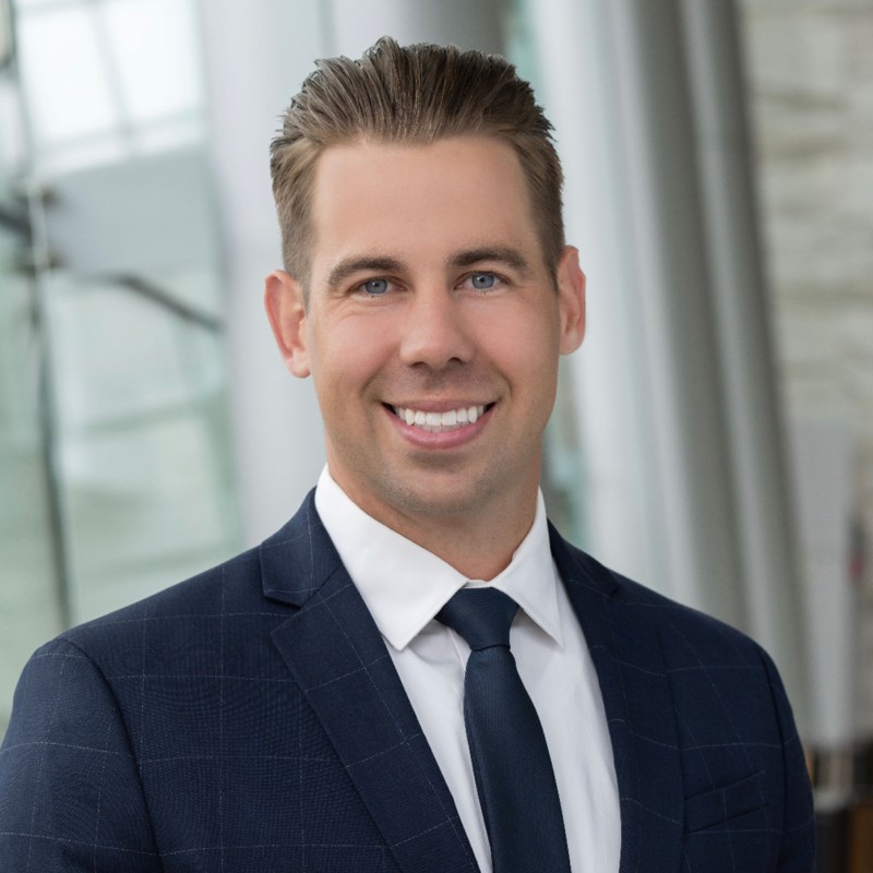 Ryan DeKimpe, Consumers Energy. A man in a navy blue suit, white shirt, and dark tie smiles at the camera. He has short, neatly styled hair and is standing indoors with a blurred modern office background.