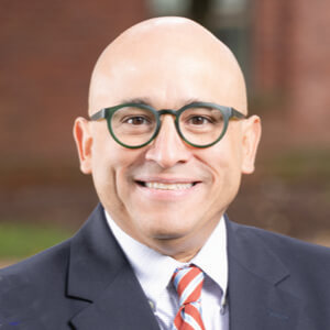 Sergio Carillo, Connecticut Green Bank. A smiling, bald man wearing round glasses, a dark suit jacket, white shirt, and red-and-blue striped tie, with a blurred outdoor background.