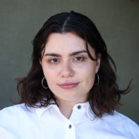 Temma Pelletier, Sunrun. A person with wavy, shoulder-length dark hair wearing a white button-up shirt and hoop earrings is facing the camera and smiling slightly. The background is plain and neutral.