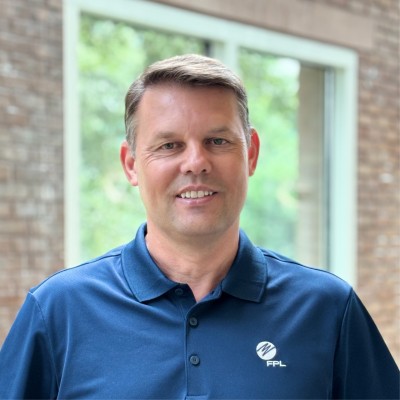 Tommy Gardner, Florida Power and Light. A man with short brown hair wearing a blue FPL polo shirt smiles while standing indoors in front of a large window with a blurred outdoor view.