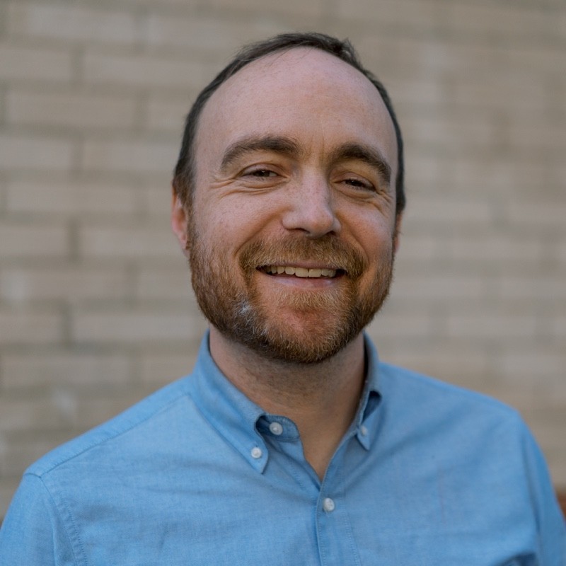 Mathias Bell, WeaveGrid. A man with short brown hair and a beard smiles at the camera. He is wearing a light blue button-up shirt and standing in front of a beige brick wall.