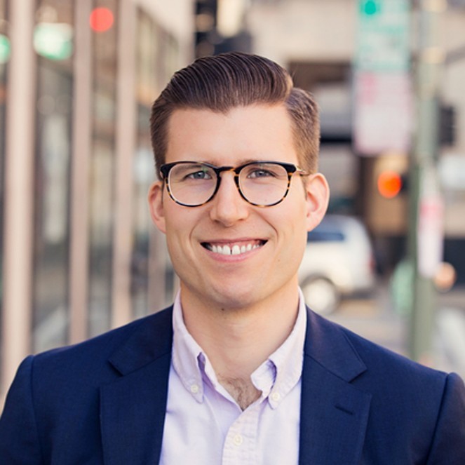 Ryan Bird, Energy Solutions. A young man with short brown hair and glasses smiles at the camera. He is wearing a navy blazer and light-colored shirt, standing on a city street with blurred cars and buildings in the background.
