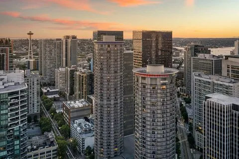 A cityscape at sunset featuring modern high-rise buildings, with the Space Needle visible in the background and a sky streaked with orange clouds.