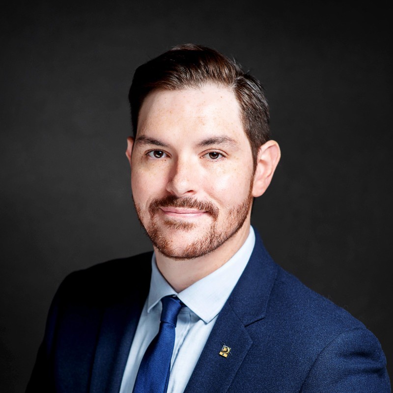 Eugene Hamrick, Rappahannock Electric Cooperative. A man with short dark hair and a trimmed beard, wearing a navy suit, light blue shirt, and blue tie, poses for a professional headshot against a dark background.