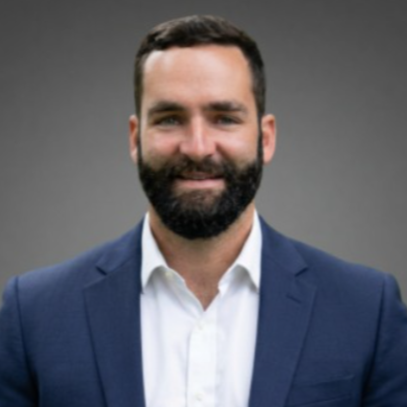 A man with short dark hair and a beard, wearing a blue suit jacket and white dress shirt, smiles in front of a plain gray background.