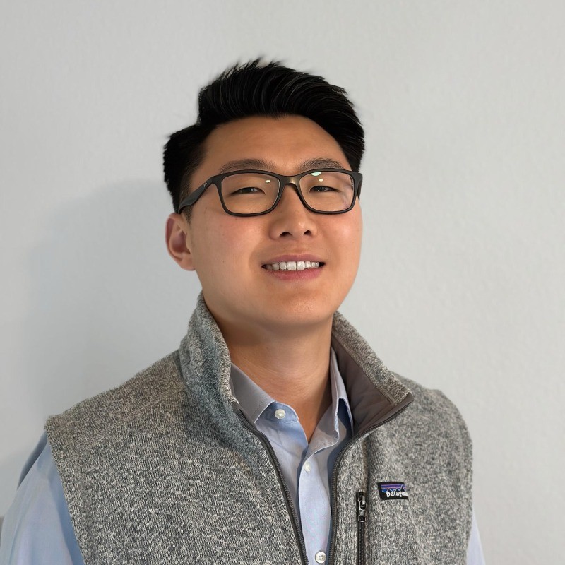 Josh Lee, Carrier. A young man with short black hair and glasses smiles at the camera. He is wearing a light blue button-down shirt under a gray fleece vest, standing in front of a plain light-colored wall.