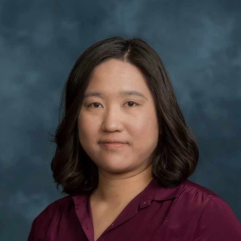 A woman with shoulder-length dark hair wearing a burgundy blouse poses for a professional headshot against a blue mottled background.