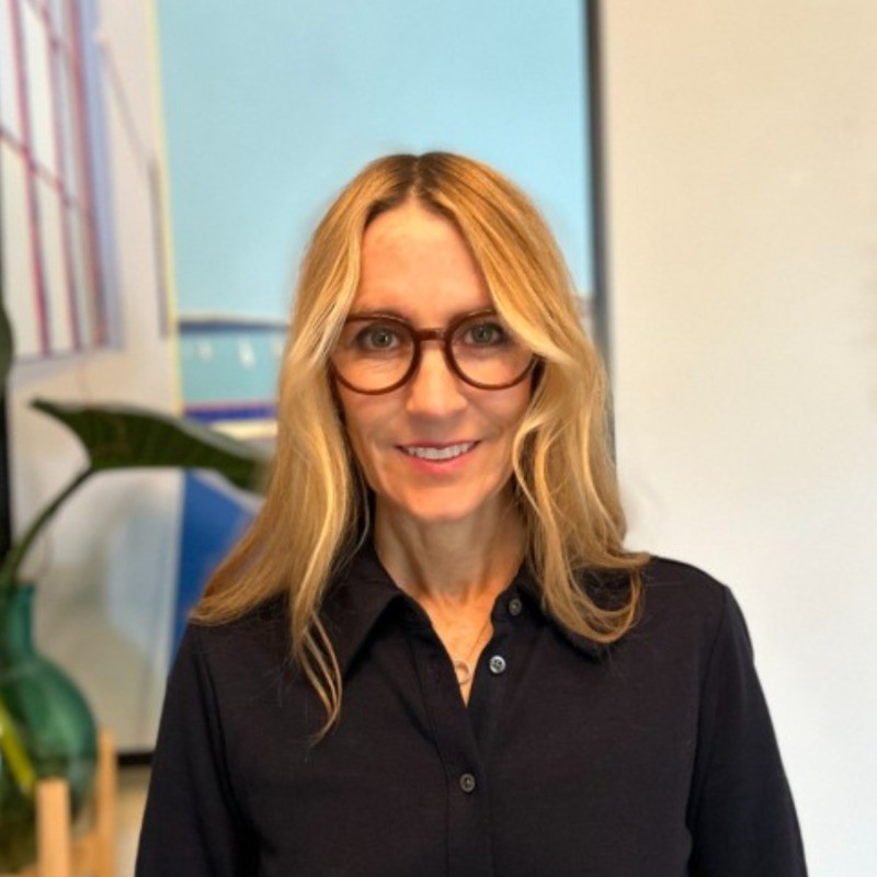 Leanne Hoadley, California Public Utilities Commission . A woman with long blonde hair and glasses, wearing a dark button-up shirt, stands indoors in front of abstract artwork and a green plant, smiling at the camera.