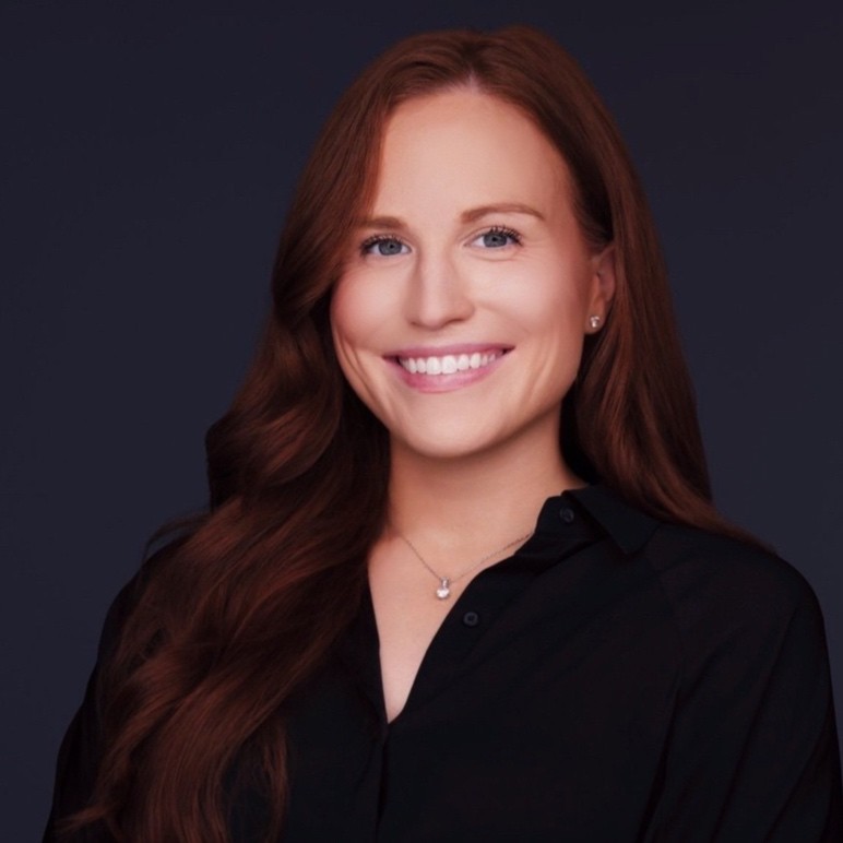 Lindsay Toth, Xcel Energy. A woman with long, wavy auburn hair smiles at the camera. She is wearing a black button-up shirt and a delicate necklace. The background is dark and plain.